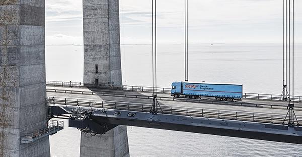 Blå lastbil kører på stor hængebro over havet med stålkabler og betontårne Blå fragtlastbil kører på en moderne hængebro med synlige stålkabler og store betontårne over roligt hav