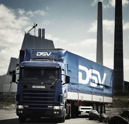 Front- og sidebillede af en blå Scania-lastbil med DSV-logo på trailer, parkeret ved et industrimiljø med høje skorstene og en skyet himmel