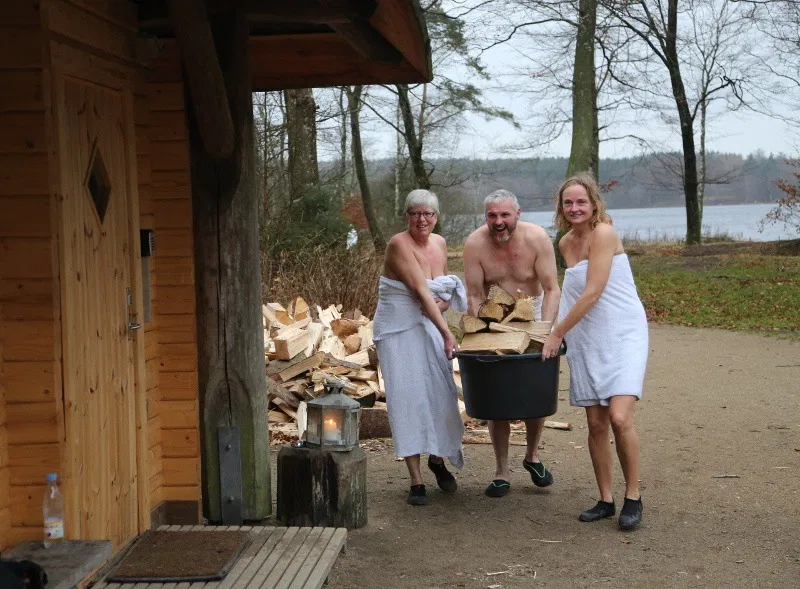 Tre smilende voksne iført håndklæder bærer en stor plastikspand fyldt med brænde udenfor en træsauna ved en sø, med en stak brænde, lanterne og træer i baggrunden