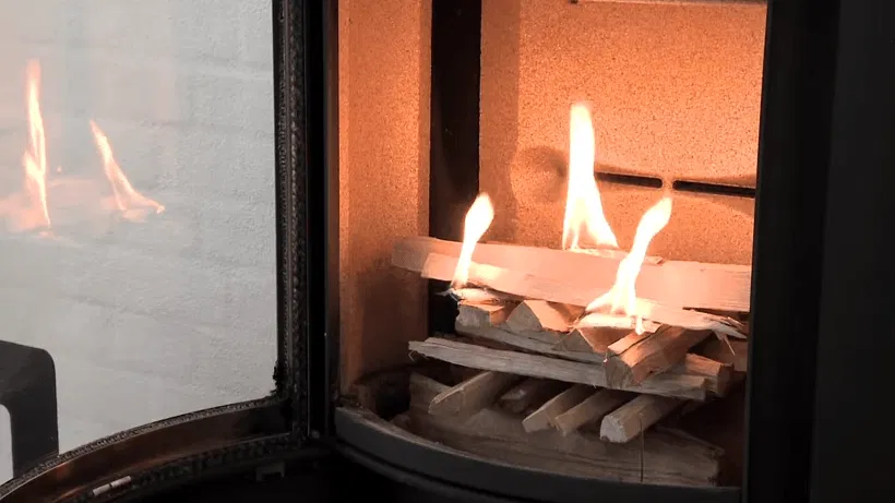 Nærbillede af tændt brændeovn med flammer, optændingsbrædder og stykke brænde bag en lukket glasdør med refleksion