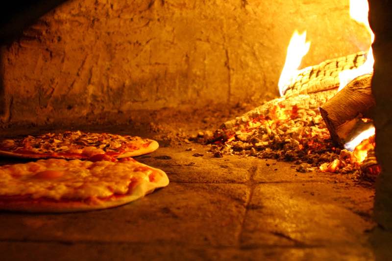 To pizzaer bager i en varm træfyret stenovn med flammer og glødende brændestykker