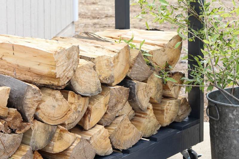 Nærbillede af stablet kløvet brænde på et sort brændestativ ved en terrasse, med en metalspand og grøn plante ved siden af