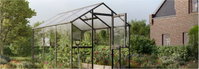 Drivhus med sort ramme og åben dør i blomstrende have Glasdrivhus med sort aluminiumsramme, åben dør, planter og en skovl i en frodig have foran et murstenshus