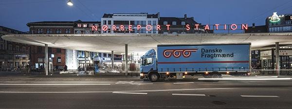 Nørreport Station i København om aftenen med blå Danske Fragtmænd-lastbil foran Aftenbillede af Nørreport Station i København med oplyst stationsskilt, perrontag og en parkeret blå Danske Fragtmænd-lastbil på gaden