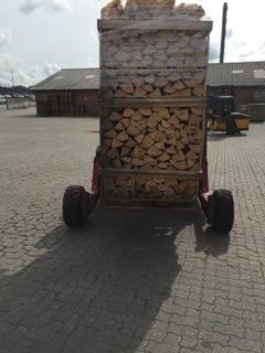 Rød trailer med stablet brænde i metalramme på gårdsplads Rød trailer lastet med stablet brænde i metalramme parkeret på brostensbelagt gårdsplads foran lave industribygninger og en palleløfter under en overskyet himmel