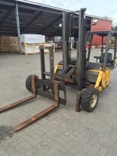 Gul brugt lille gaffeltruck med to gafler og pneumatiske dæk parkeret udendørs ved lager Gul gaffeltruck i brugt stand med to gafler, mast og pneumatiske dæk, parkeret på brolagt område foran lagerbygninger